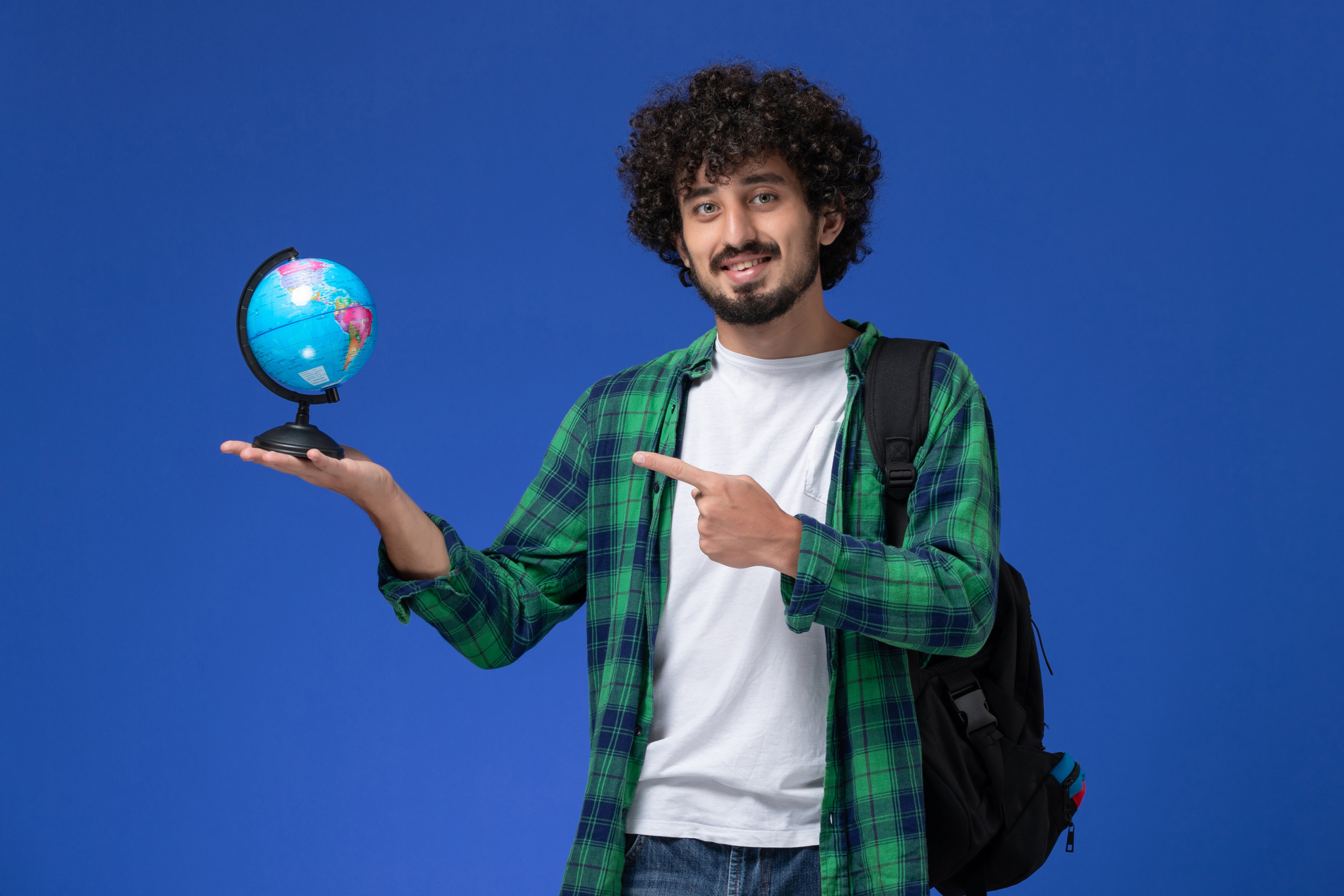 front-view-male-student-green-checkered-shirt-wearing-black-backpack-holding-little-globe-smiling-blue-wall